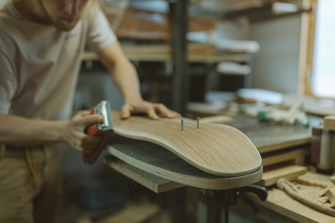 A craftsman carefully sands a wooden skateboard deck in a workshop setting, focused on precision and detail.